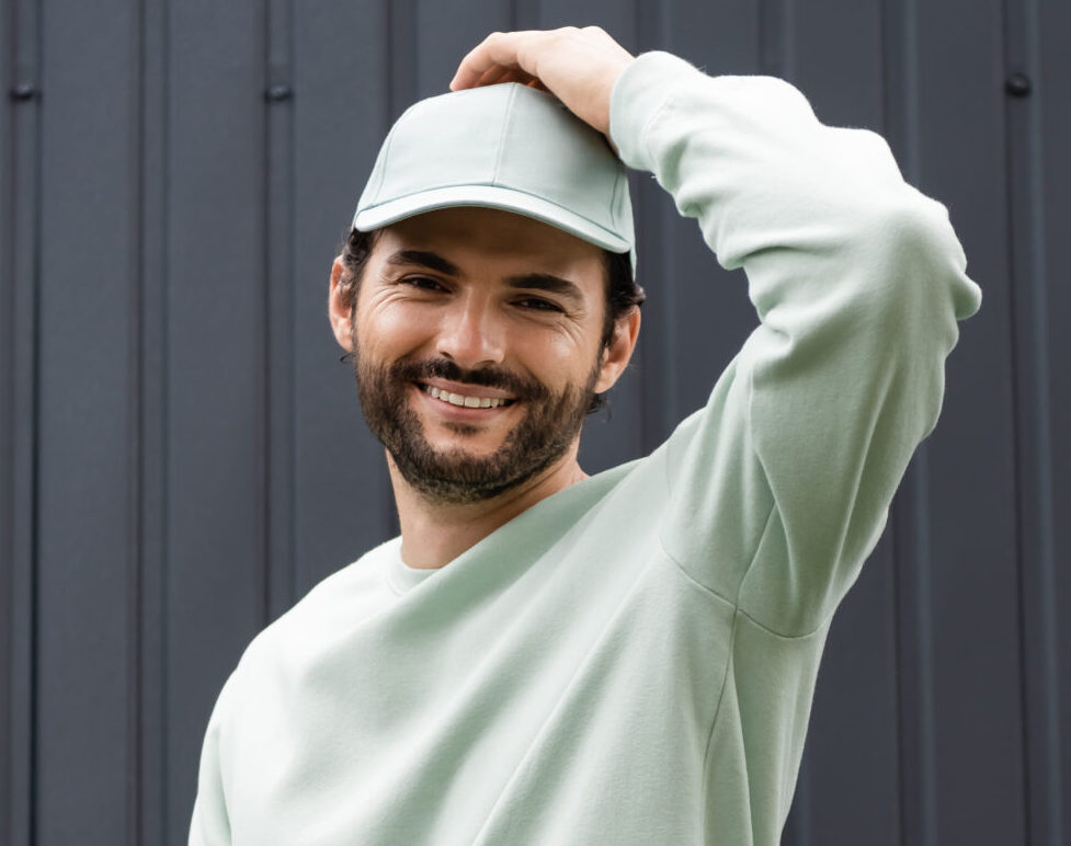 smiling man in sweatshirt adjusting cap near metallic fence, banner